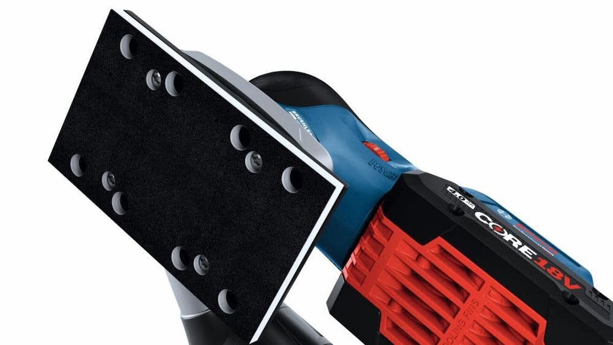 An inverted view focusing on the undercarriage of the Bosch 18V finishing sander displaying a black hook-and-loop rectangular sanding plate with built-in dust extraction vacuum holes alongside a mounted red and black battery pack.