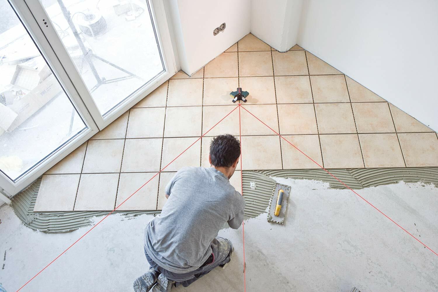 A tiler kneels to spread mortar, following the 45-degree and 90-degree red laser lines projected by the Bosch GTL3 for a diagonal floor tile layout.