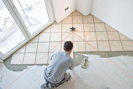 A tiler kneels to spread mortar, following the 45-degree and 90-degree red laser lines projected by the Bosch GTL3 for a diagonal floor tile layout.