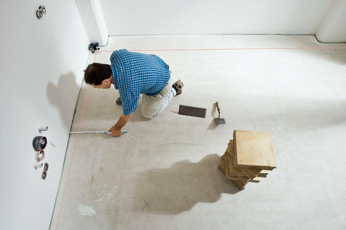 A tiler kneels on a concrete floor to take measurements, using the Bosch GTL3's projected 90-degree red laser lines for a square layout.