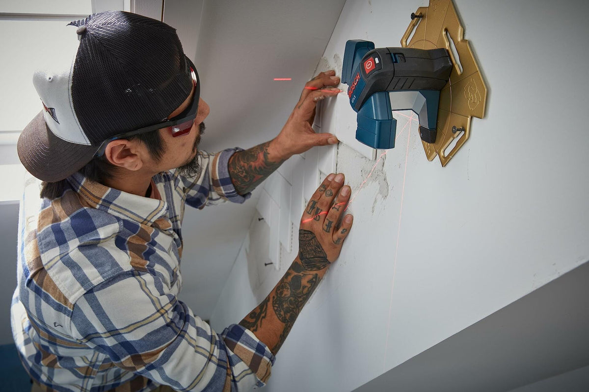 A worker mounts the Bosch GTL3 tile laser onto its wall positioning plate, projecting red vertical and horizontal lines for layout.