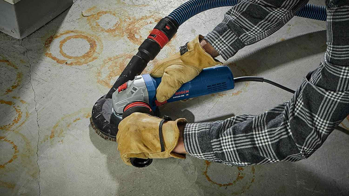 A worker using the angle grinder equipped with a dust shroud attachment and connected to a vacuum hose to grind a concrete floor.