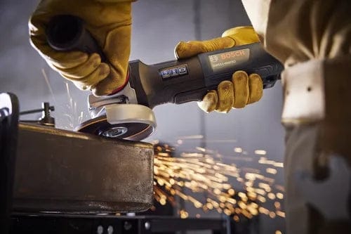A close-up action shot of a person wearing protective yellow gloves using the Bosch angle grinder to grind the edge of a thick metal plate, with bright sparks flying from the contact point.