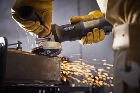 A close-up action shot of a person wearing protective yellow gloves using the Bosch angle grinder to grind the edge of a thick metal plate, with bright sparks flying from the contact point.
