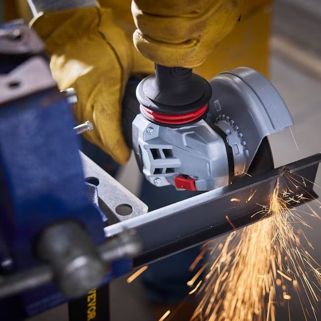 An operator wearing yellow protective gloves uses the Bosch 5-inch angle grinder to cut through a metal L-bracket secured in a blue vise, demonstrating heavy-duty metal cutting capabilities with sparks flying.