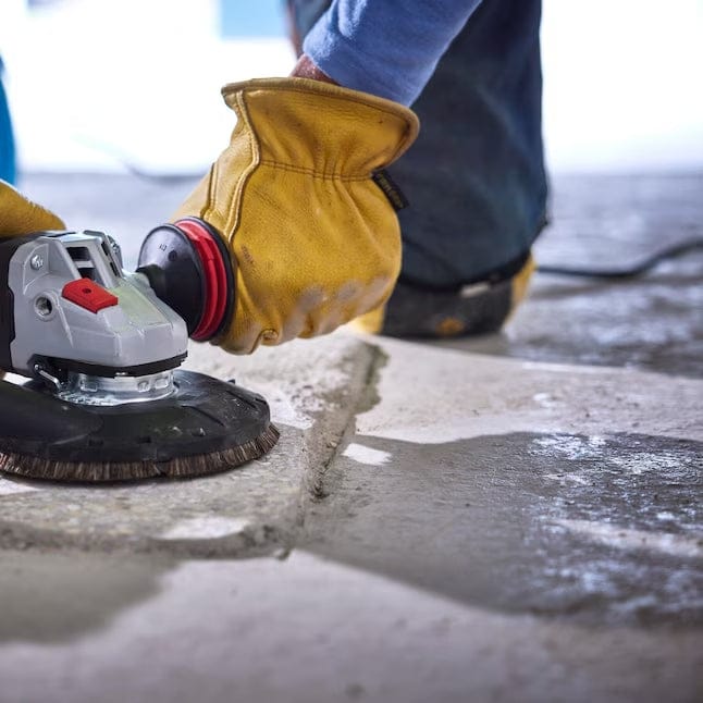 An action shot showing an operator in yellow protective gloves using the Bosch 6-inch angle grinder with a specialized surface grinding shroud and brush skirt to smooth a concrete slab.