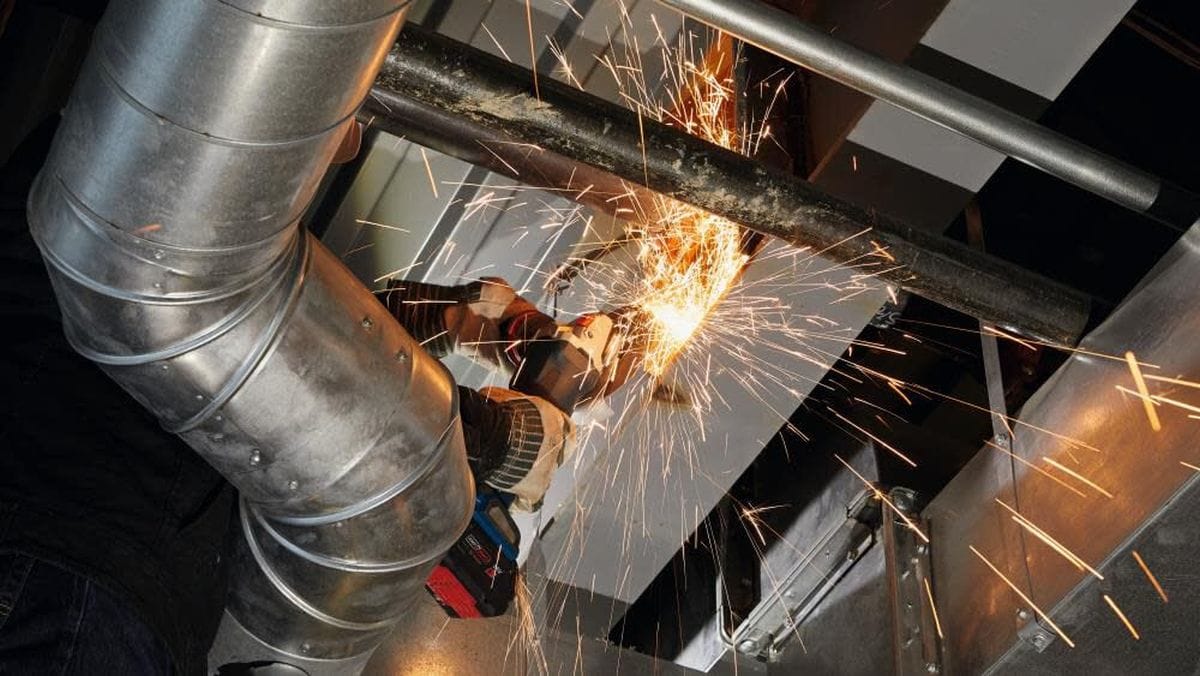 A jobsite application shot showing a professional using the Bosch cordless angle grinder to cut overhead metal piping, highlighting the tool's balance and the safety of the cordless BITURBO system.