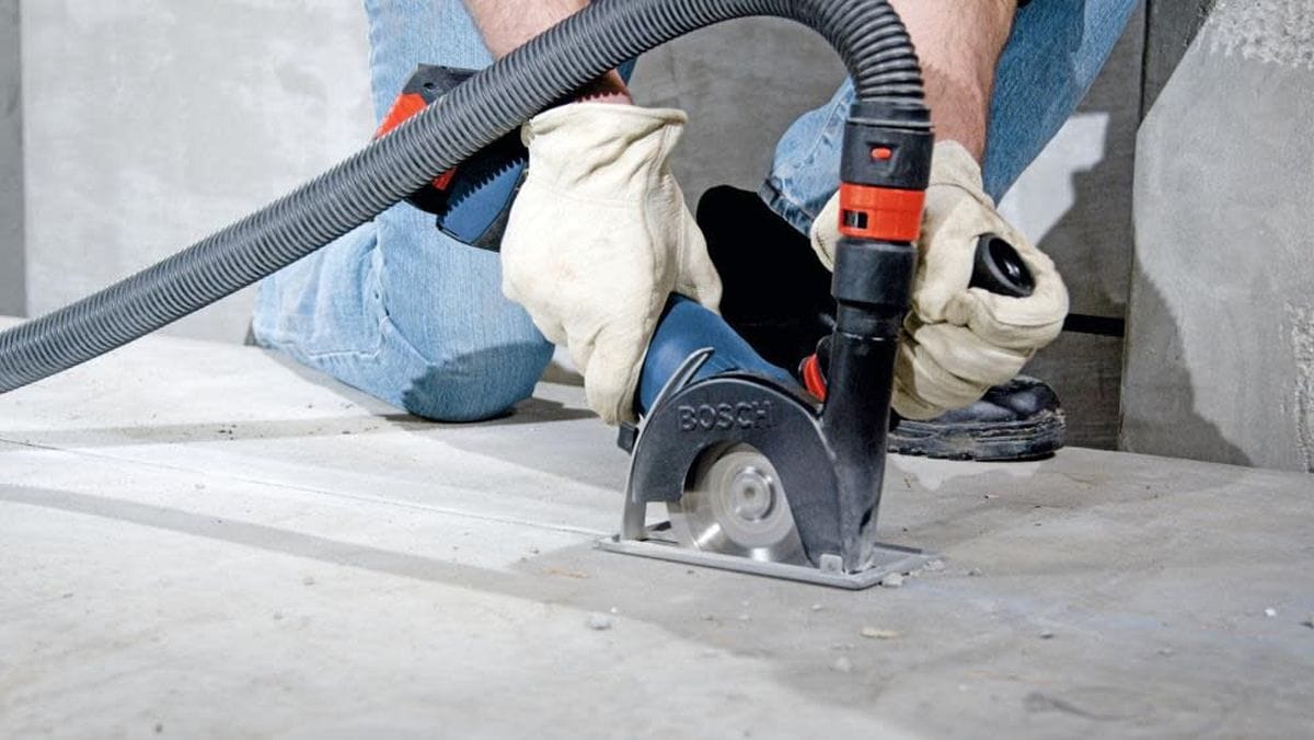 A construction professional using the Bosch 18V cordless grinder with a dust extraction shroud and vacuum attachment to perform precision concrete cutting on a jobsite floor.