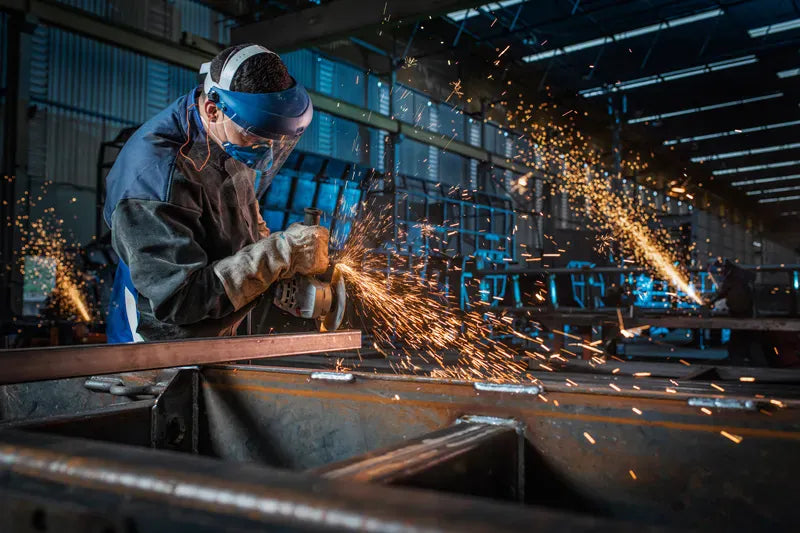 An active landscape action view identical to file GWS25-180PR 5.webp showing a fabricator running a surface disc along a carbon steel beam with the Bosch GWS25-180PR corded large angle grinder. Dropping all high-conversion text blocks, the raw shot captures a dense shower of metal sparks reflecting off the tool's impact-resistant blue resin composite body shell.