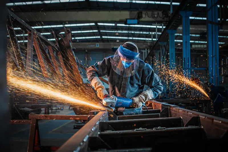 An industrial fabricator wearing clear protective safety glasses and heavy gray work gloves using the Bosch GWS25-180PR 7-inch angle grinder to slice down a vertical metal substrate. This uncropped perspective view captures a dense shower of bright orange sparks discharging backward from the closed metal cutting wheel safety guard inside a commercial workshop.
