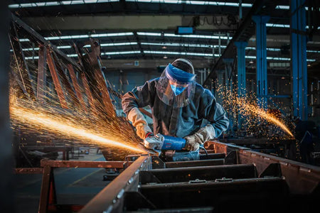 An industrial fabricator wearing clear protective safety glasses and heavy gray work gloves using the Bosch GWS25-180PR 7-inch angle grinder to slice down a vertical metal substrate. This uncropped perspective view captures a dense shower of bright orange sparks discharging backward from the closed metal cutting wheel safety guard inside a commercial workshop.