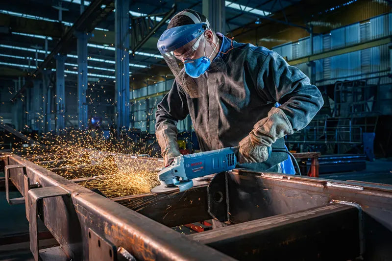 A close-up field action shot of a structural welder utilizing the Bosch GWS25-180PR 15-Amp corded large angle grinder to bevel a thick carbon steel plate. The technician grips the dual-position auxiliary handle firmly with both hands to maintain flat, even tracking control while executing heavy surface disc grinding.