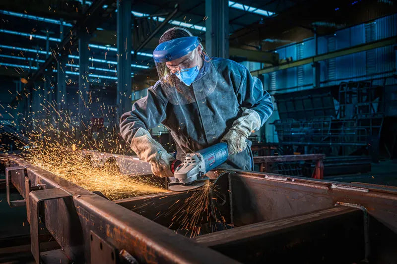 An uncropped workshop view identical to file GWS25-180PR 4.webp, capturing a professional welder smoothing out a metal union seam with the Bosch GWS25-180PR 7-inch angle grinder. This variant features no text blocks, providing a clear perspective of the tool's vibration control side grip and adjustable guard positions under ambient body shop lighting.