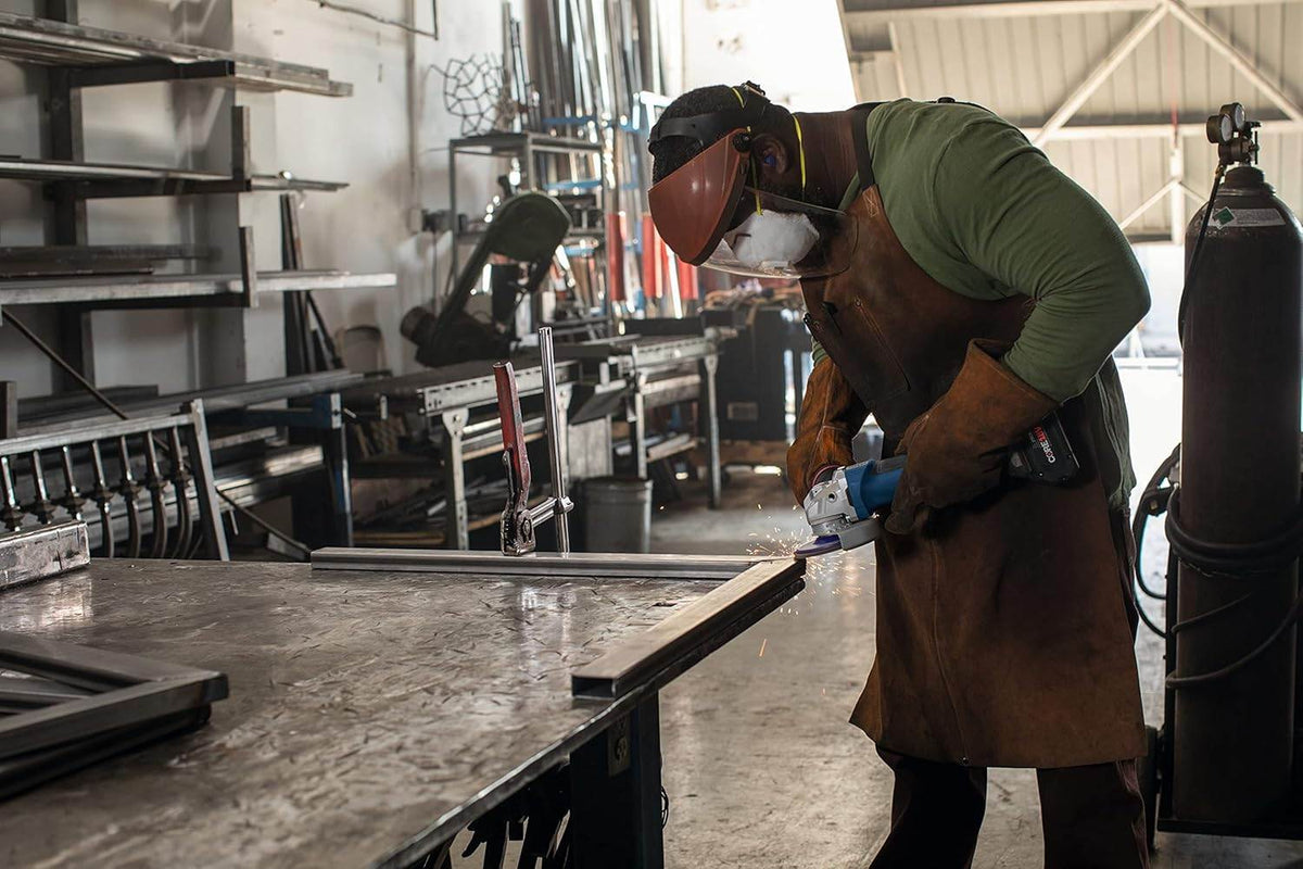 Person wearing full protective gear using angle grinder on metal piece; sparks flying in industrial workshop setting.