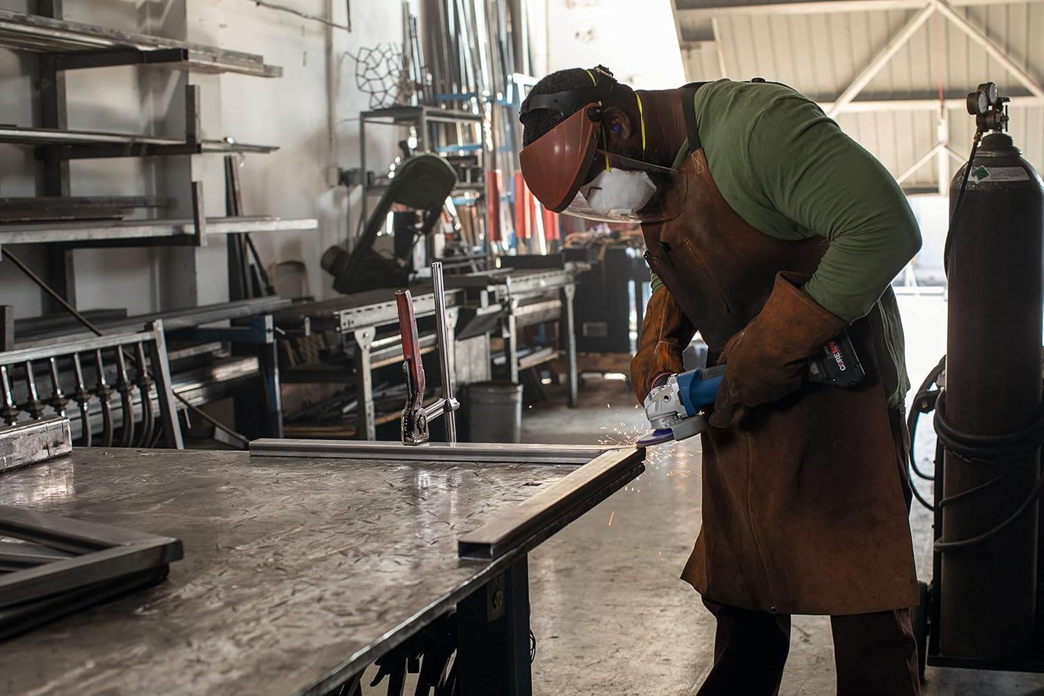 Person wearing full protective gear using angle grinder on metal piece; sparks flying in industrial workshop setting.