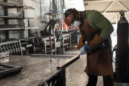 Person wearing full protective gear using angle grinder on metal piece; sparks flying in industrial workshop setting.