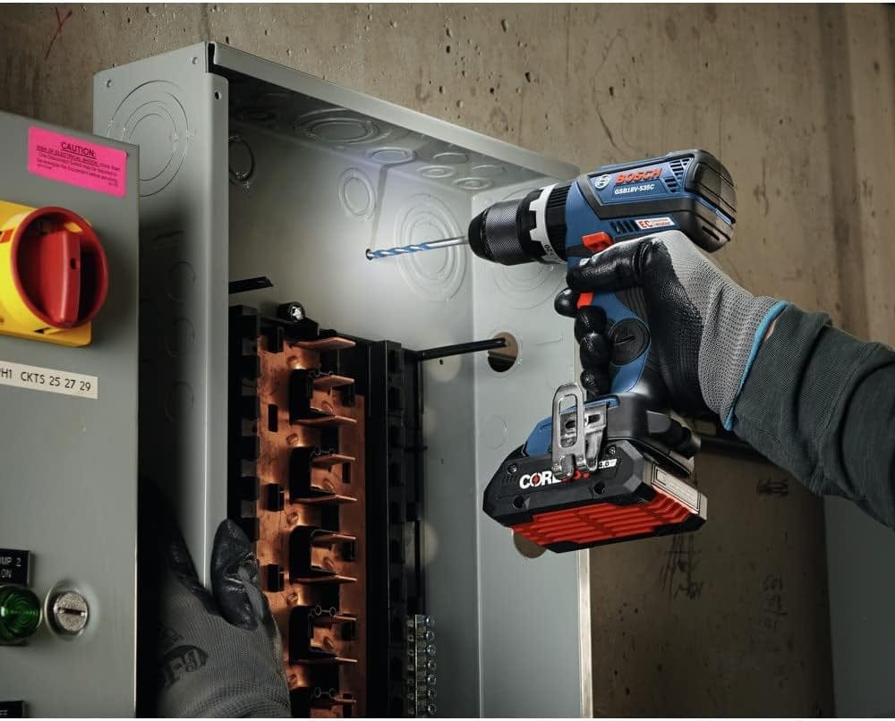A professional using the Bosch 18V hammer drill to drill into a metal electrical panel box on a jobsite, showcasing the integrated LED light and precision control.