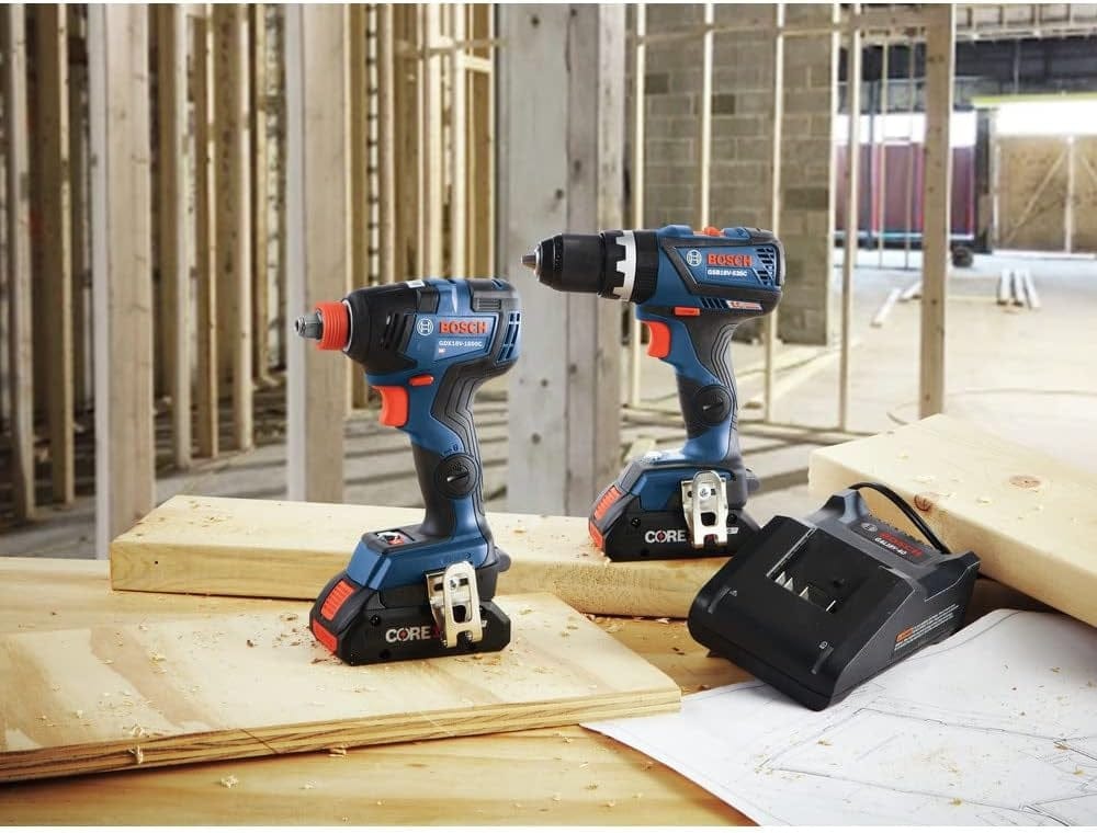 A jobsite display of the Bosch 18V brushless hammer drill, Freak impact driver, and battery charger on wood planks at a construction site to demonstrate professional-grade tool availability.