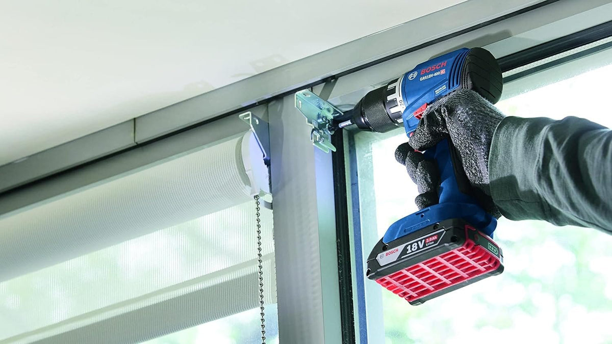 A gloved hand operating the Bosch 18V brushless drill driver to fasten metal hardware above a window frame, demonstrating how the built-in LED work light clearly illuminates shadowed corners for accurate installation.