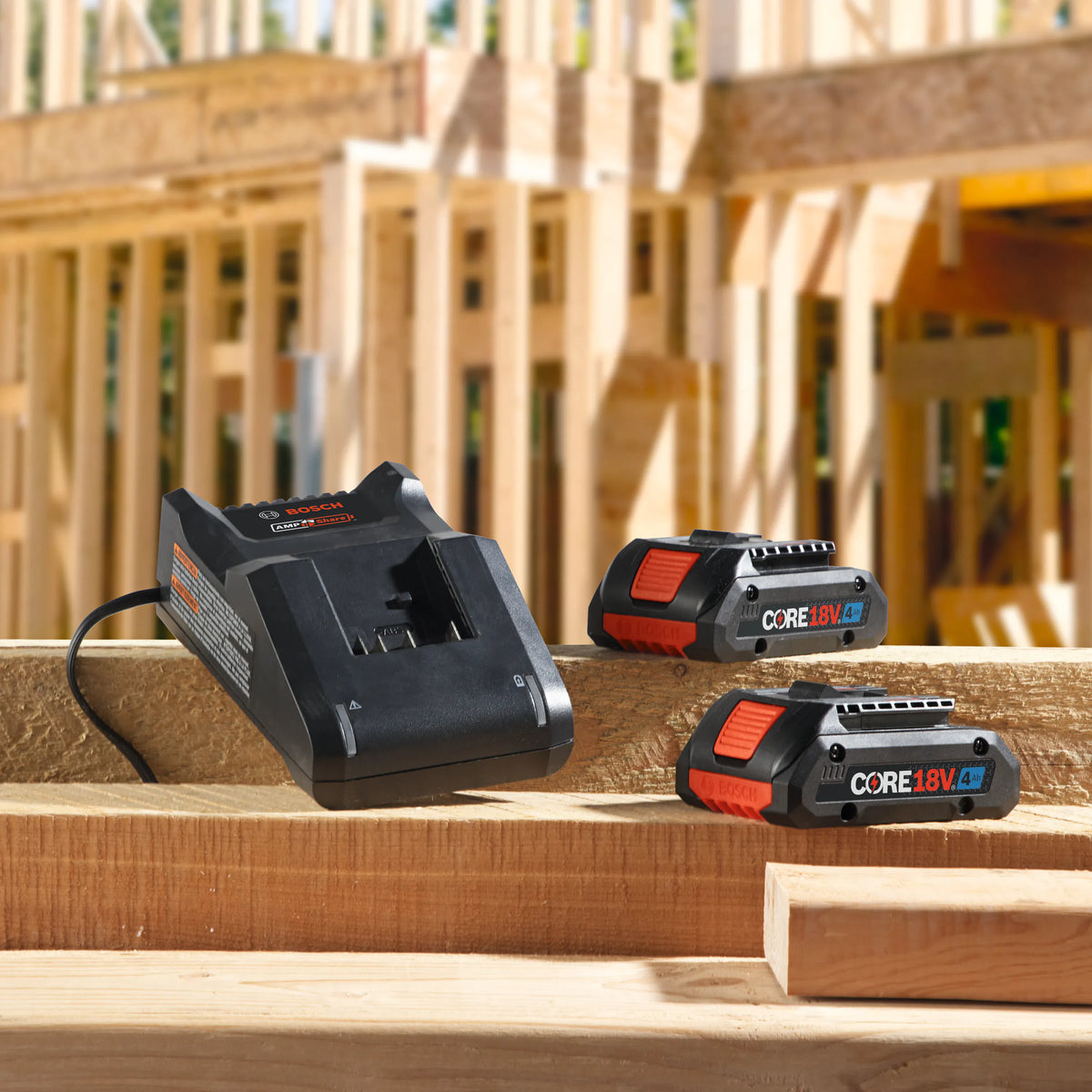 A jobsite lifestyle photograph of the Bosch 18V starter kit, displaying the GAL18V-40 fast charger docked next to two CORE18V 4.0 Ah compact batteries resting over a raw wooden framing beam at a residential home building project.