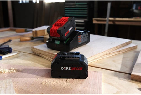 Bosch 18V battery and charger kit positioned on a wooden jobsite surface surrounded by wood shavings and clamps.