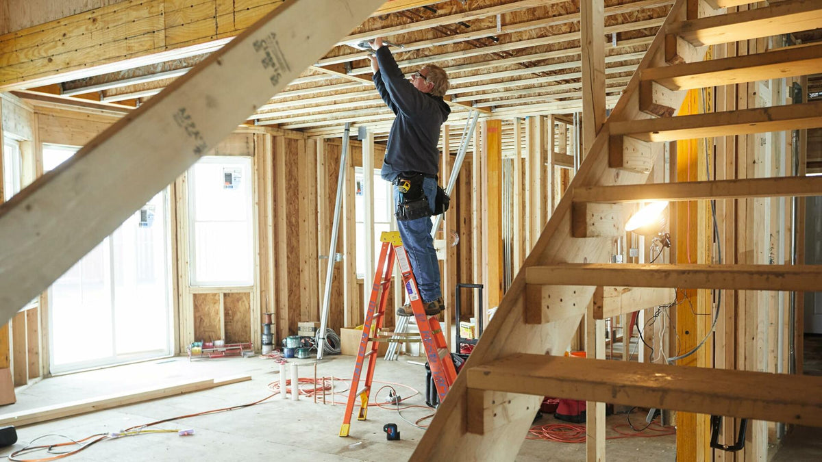 Construction worker standing on a step ladder inside a framed building, performing overhead installation or layout work, demonstrating the plumb function of the laser