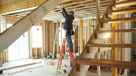 Construction worker standing on a step ladder inside a framed building, performing overhead installation or layout work, demonstrating the plumb function of the laser