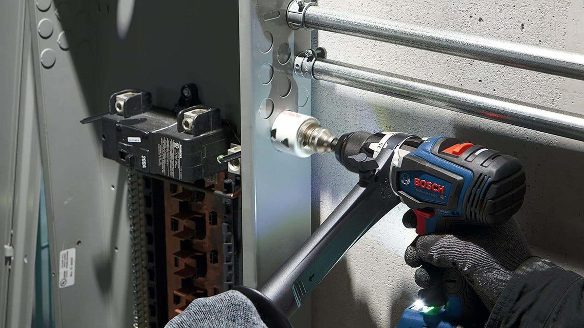 A worker uses a Bosch drill and the hole saw to cut through the side of a grey metal electrical panel box.