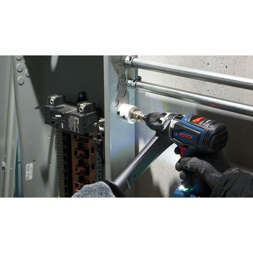 A gloved hand uses a Bosch drill with an auxiliary handle and a white bi-metal hole saw to drill a hole in the side of a metal electrical panel.