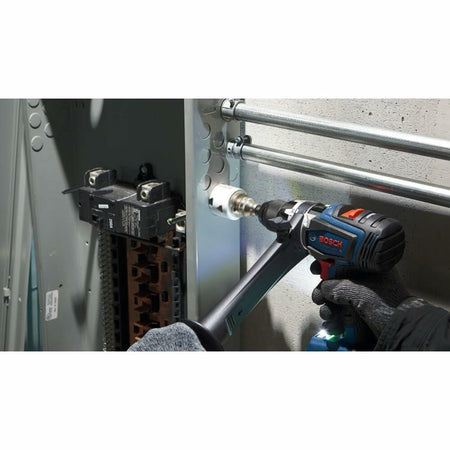 A gloved hand uses a Bosch drill with an auxiliary handle and a white bi-metal hole saw to drill a hole in the side of a metal electrical panel.