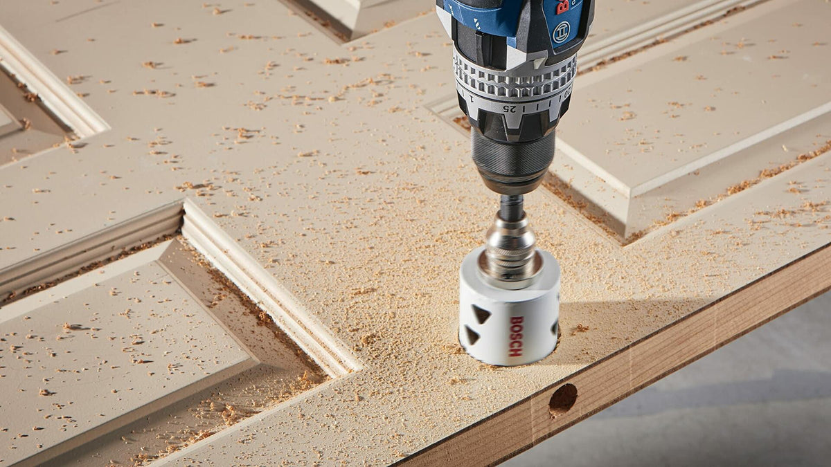 A Bosch drill with a white bi-metal hole saw is shown drilling a hole for a doorknob in a white, six-panel wooden door.
