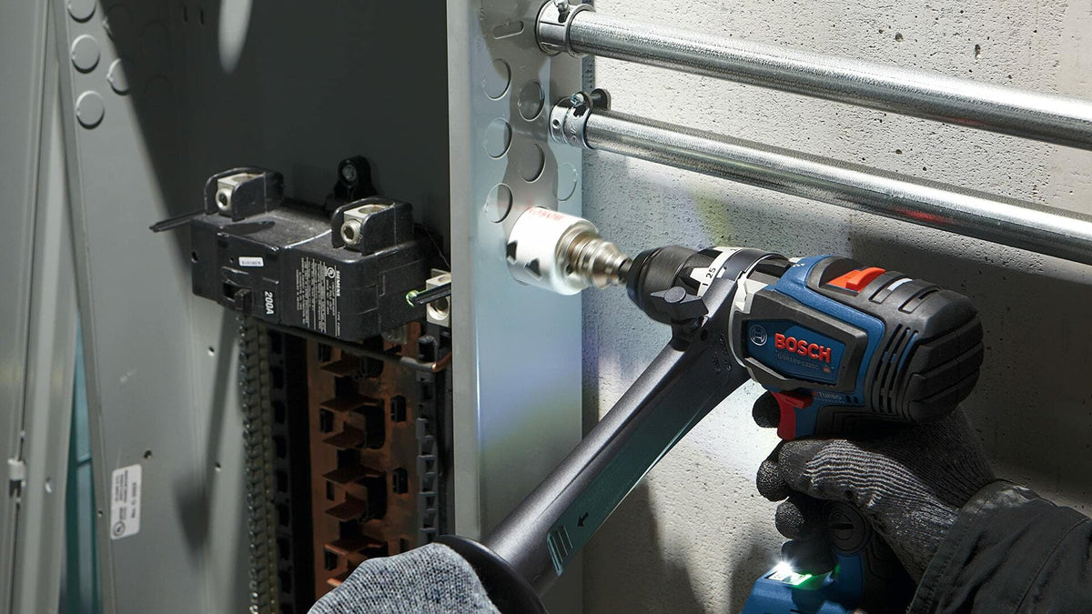 A gloved hand uses a Bosch drill with an auxiliary handle and a white bi-metal hole saw to drill a hole in the side of a metal electrical panel.