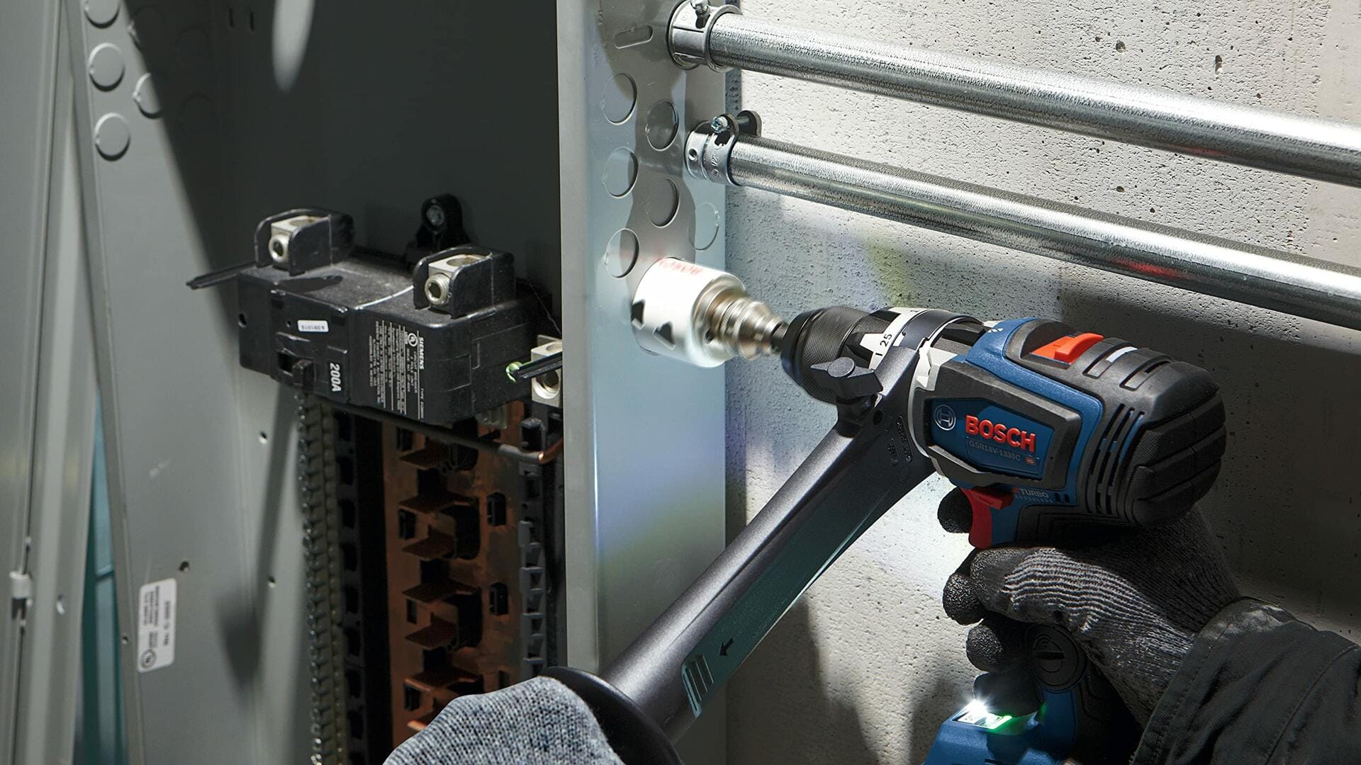 A gloved hand uses a Bosch drill with an auxiliary handle and a white bi-metal hole saw to drill a hole in the side of a metal electrical panel.