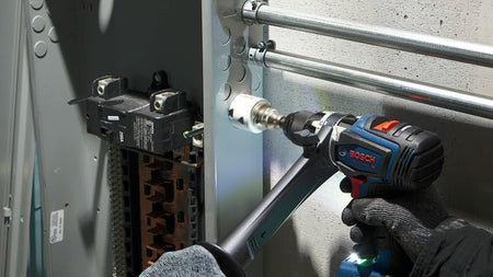 A gloved hand uses a Bosch drill with an auxiliary handle and a white bi-metal hole saw to drill a hole in the side of a metal electrical panel.