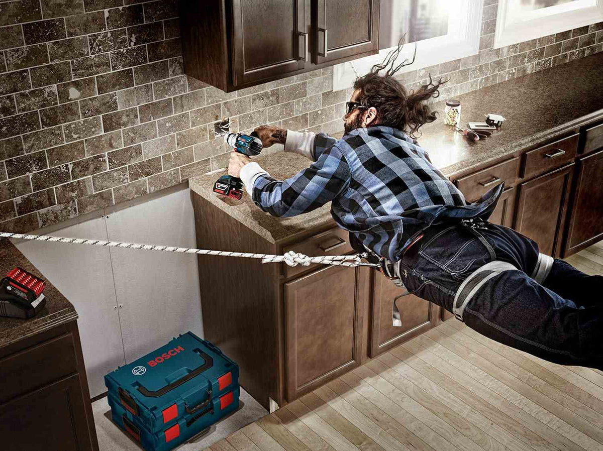 A promotional action shot showing a worker suspended by a rope harness while using a hammer drill and BlueGranite bit to drill into a kitchen backsplash.