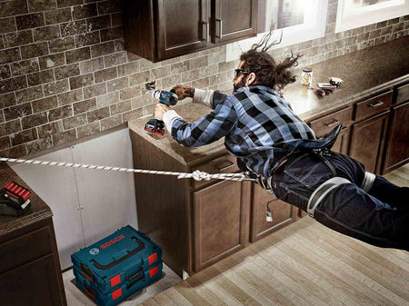 A promotional action shot showing a worker suspended by a rope harness while using a hammer drill and BlueGranite bit to drill into a kitchen backsplash.