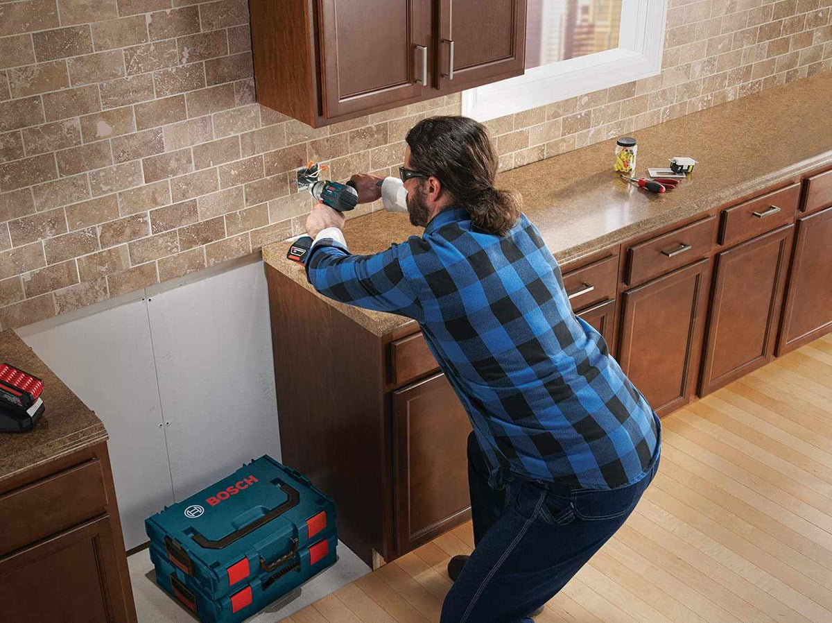 A worker in a plaid shirt uses a cordless hammer drill with the BlueGranite bit to drill into a stone tile backsplash in a kitchen.