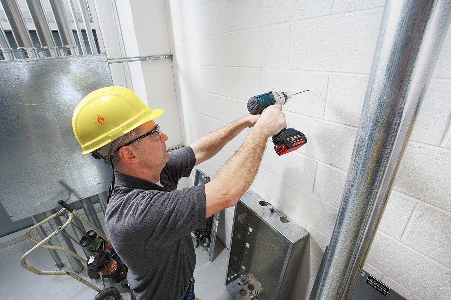 A worker wearing a yellow hard hat and safety glasses uses a hammer drill to drill into a white cinder block wall in an industrial setting.