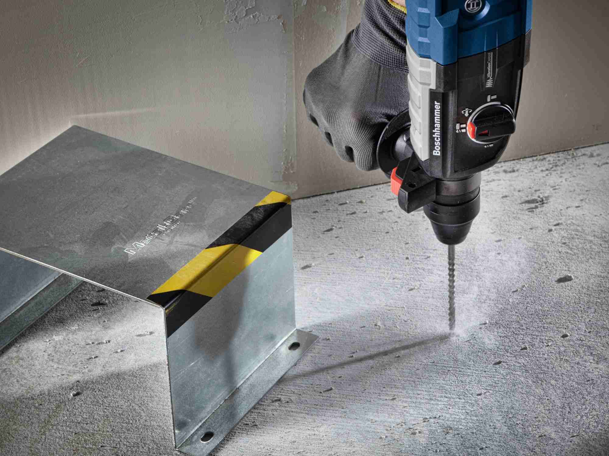 An action shot of a professional user operating a Bosch rotary hammer to drill a vertical hole into a concrete floor, demonstrating the bit's precision near metal fixtures.