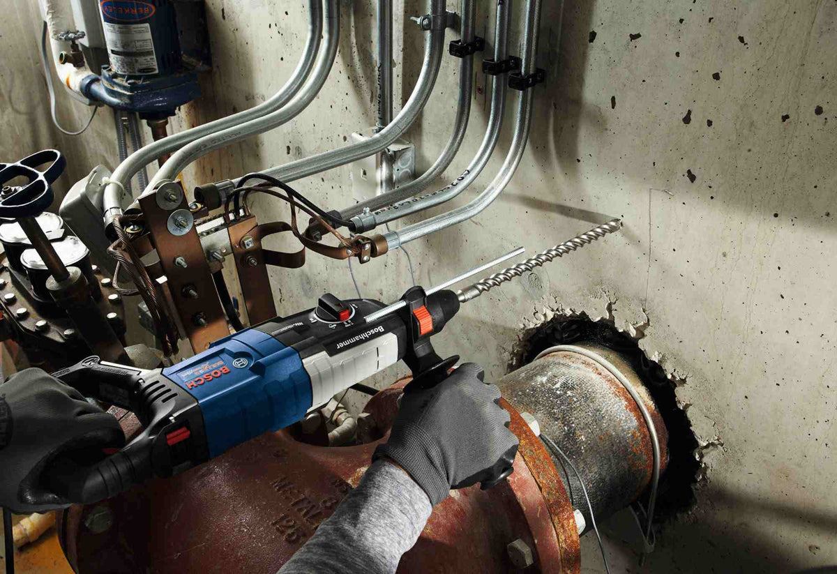 A wide action shot of a professional using a Bosch rotary hammer and a long Bulldog Xtreme bit to drill horizontally into a concrete wall in a mechanical room with exposed piping.