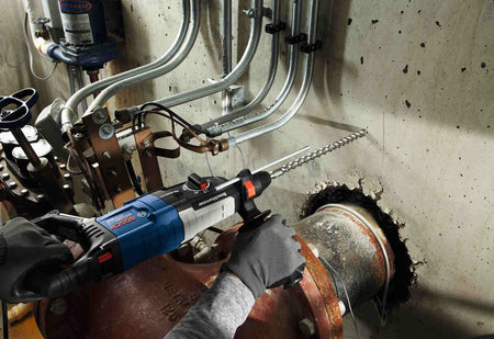 A wide action shot of a professional using a Bosch rotary hammer and a long Bulldog Xtreme bit to drill horizontally into a concrete wall in a mechanical room with exposed piping.