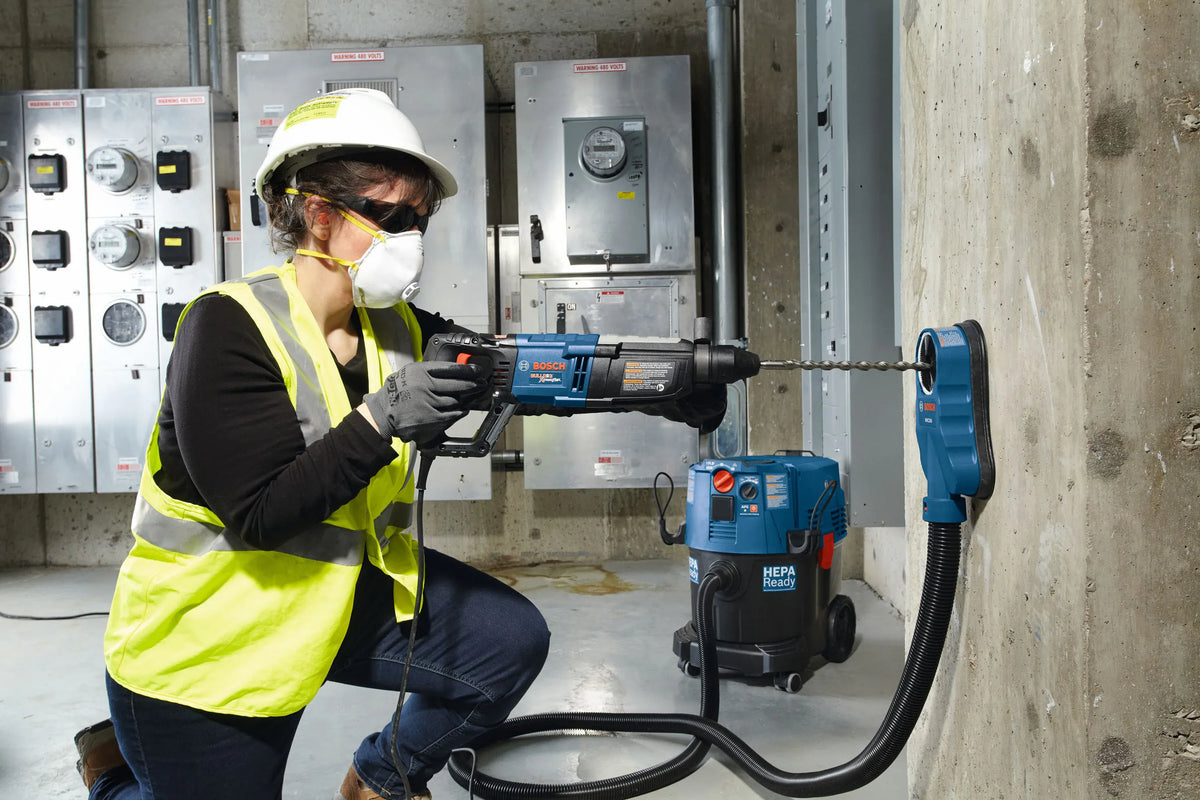 A female construction worker wearing a hard hat, safety glasses, face mask, and high-visibility vest kneels while using a Bosch corded rotary hammer drill equipped with a long carbide masonry drill bit from the HCK001 set. She is drilling into a concrete column with an attached dust extraction shroud connected to a blue HEPA-ready jobsite vacuum.