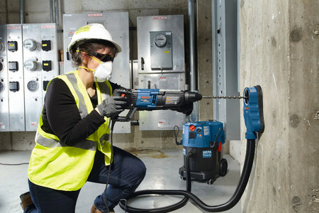 A female construction worker wearing a hard hat, safety glasses, face mask, and high-visibility vest kneels while using a Bosch corded rotary hammer drill equipped with a long carbide masonry drill bit from the HCK001 set. She is drilling into a concrete column with an attached dust extraction shroud connected to a blue HEPA-ready jobsite vacuum.