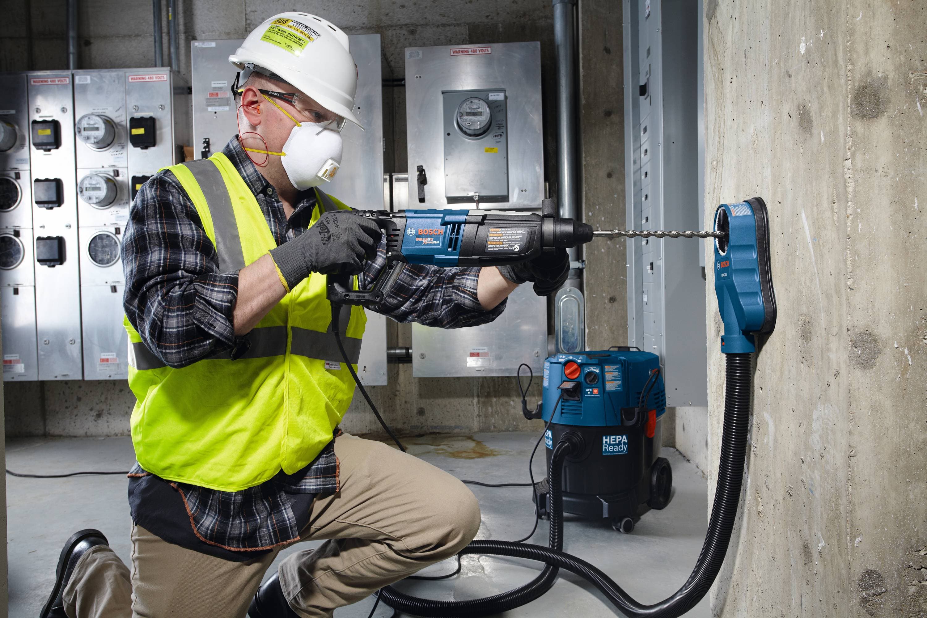 A male tradesperson wearing full personal protective equipment, including a hard hat, respirator mask, safety glasses, and high-visibility vest, operates a professional Bosch Bulldog SDS Plus rotary hammer drill in an industrial electrical room. He uses a heavy duty masonry bit from the Bosch HCK001 7 piece set to drill into a solid concrete wall utilizing a dust extraction vacuum system.