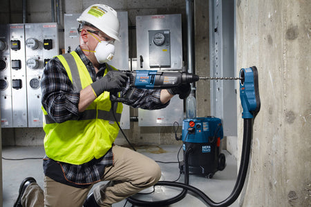 A male tradesperson wearing full personal protective equipment, including a hard hat, respirator mask, safety glasses, and high-visibility vest, operates a professional Bosch Bulldog SDS Plus rotary hammer drill in an industrial electrical room. He uses a heavy duty masonry bit from the Bosch HCK001 7 piece set to drill into a solid concrete wall utilizing a dust extraction vacuum system.