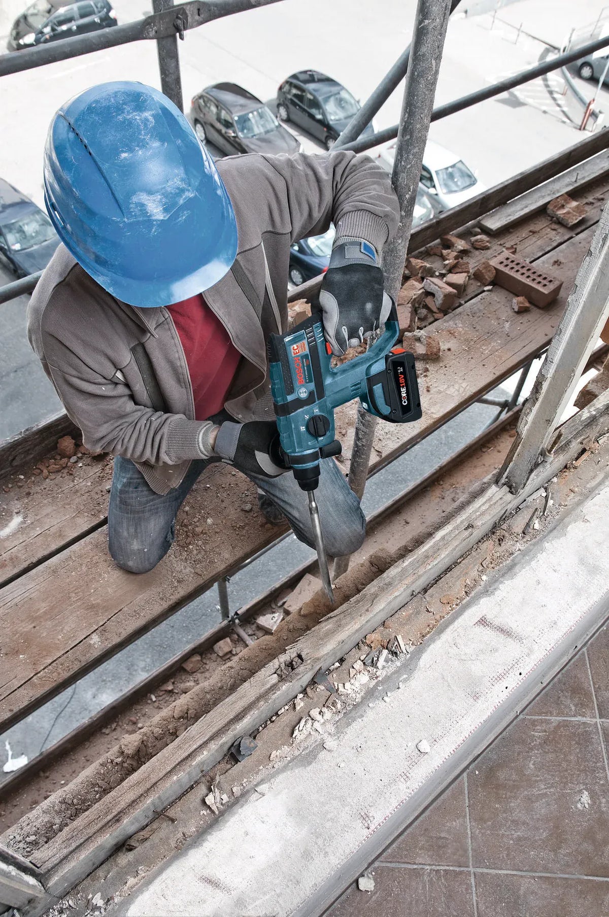 A high-angle view of a trade professional wearing a blue hard hat, heavy work gloves, and protective gear while kneeling on a high scaffolding structure. The worker is operating a Bosch cordless SDS Plus rotary hammer drill equipped with a long point chisel to break down and clear old brickwork and concrete debris.
