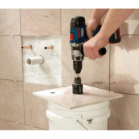 Action shot of a professional using a cordless drill equipped with the Bosch HDG4 diamond hole saw to wet-cut a hole in a ceramic tile resting on a white bucket, demonstrating the tool's use for bathroom plumbing installations.