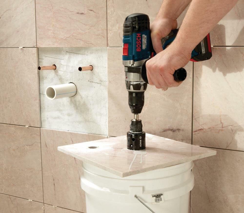 A person uses a Bosch drill with a diamond hole saw to cut a hole in a tile, which is resting on top of a bucket for support in a partially tiled bathroom.