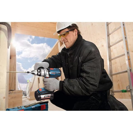 Worker driving screw into wooden frame with Bosch 18V cordless drill; safety gear and toolbox visible on jobsite.