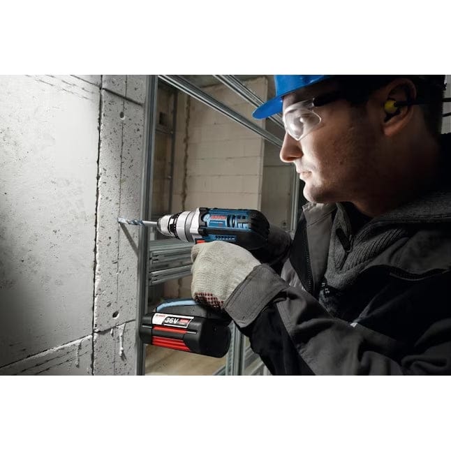 Person using Bosch cordless hammer drill to bore into concrete wall; wearing full safety gear on construction site.